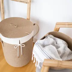 Naturalny pojemnik na drewno z juty, z białym wykończeniem i sznurkiem. Obok kosz z ręcznikiem.